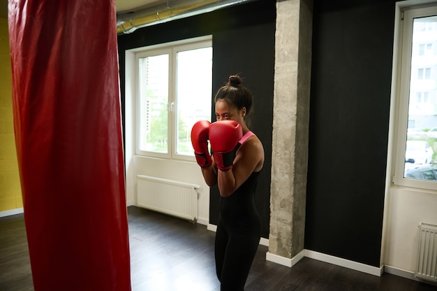 Boxer fitness donna in abbigliamento sportivo nero attillato e guantoni da boxe rossi che si allenano in una palestra di boxe, facendo un pugno dritto colpendo un sacco da boxe durante un allenamento indoor