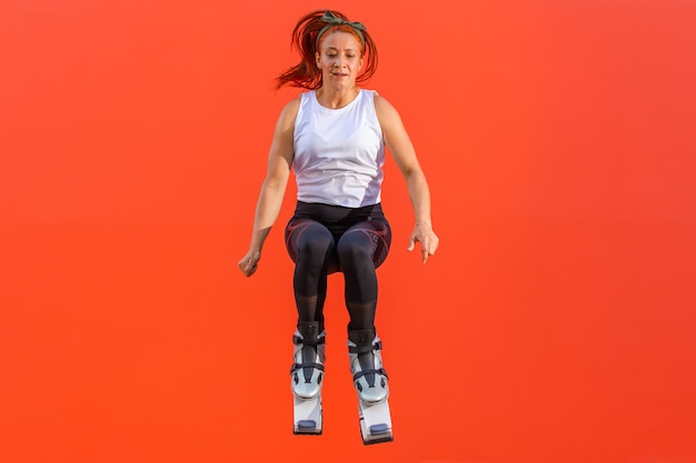 Fitness woman doing a jump wearing kangoo jumps boots on an\
orange background. concept of healthy life and sport