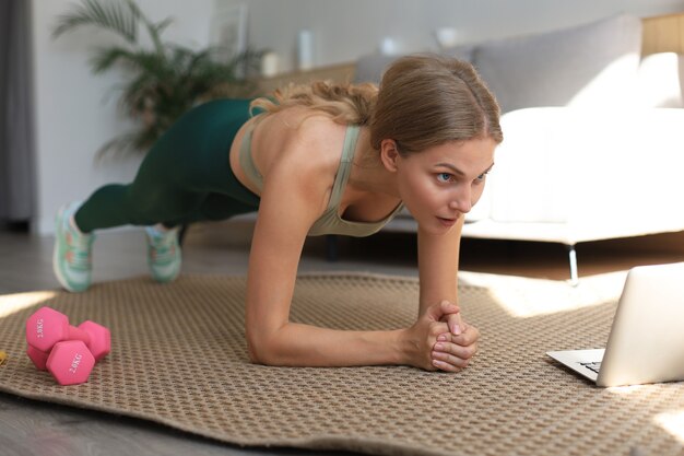 Fitness woman doing plank and watching online tutorials on laptop, training in living room. Stay at home activities.