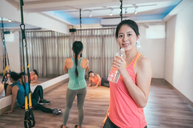 Fitness woman drinking water from bottle. muscular young female\
at gym taking a break from workout. group of tired friends sweating\
resting in the background after trx lesson at health club.