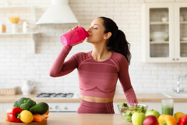 Fitness woman drinking water from bottle standing near kitchen table