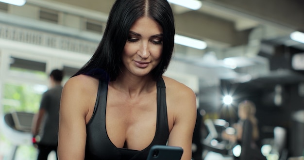 Fitness woman holding smartphone in sport club. Closeup beautiful female athlete texting message on cellphone at gym. Smiling sport woman resting with mobile phone.