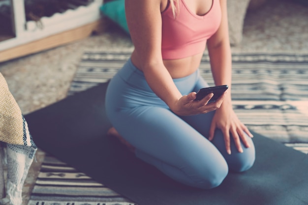 요가 매트에 무릎을 꿇고 휴대폰을 사용하는 피트니스 여성, 집에서 운동을 하는 동안 휴대폰을 사용하는 여성. 집에서 매트에 앉아 운동하는 동안 온라인 튜토리얼을 보고 있는 스포츠웨어를 입은 여성