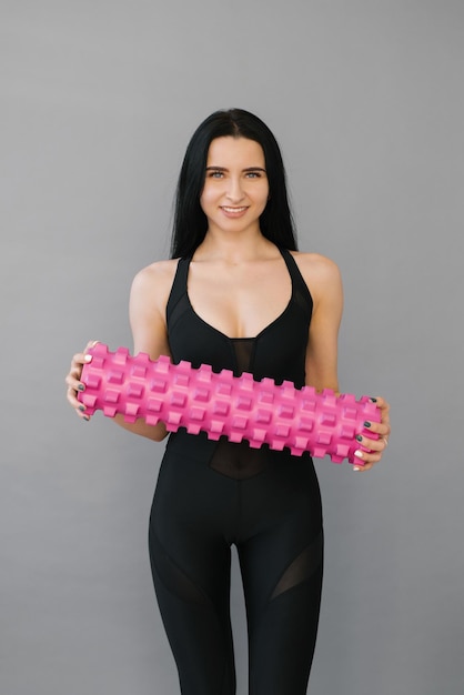 Fitness a woman in sports black clothes with a massage roller or a roll in her hands smiles on a gray background Fitness trainer with items for myofascial release