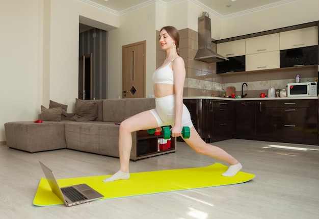 Fitness woman in sportswear does exercise with dumbbells looks into the laptop screen watches a fitness tutorial on the mat in the living room Sports at home