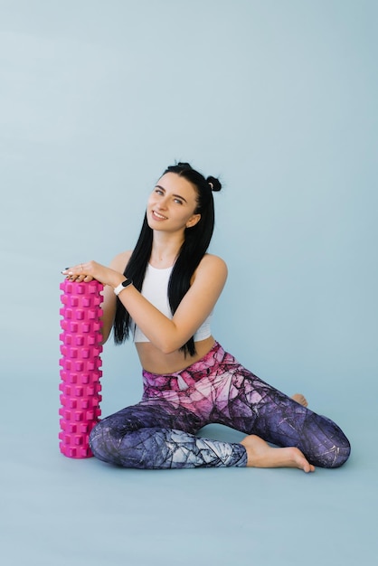 Fitness a woman in sportswear with a massage roller or a roll in her hands smiles while sitting on a blue isolated background Fitness trainer with items for myofascial release