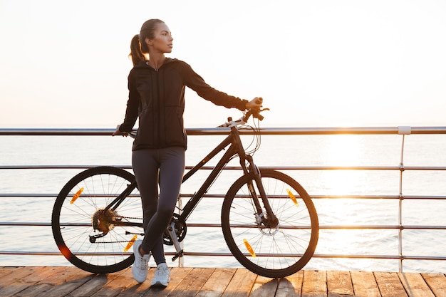 fitness woman standing with bicycle on boardwalk, during sunrise over sea