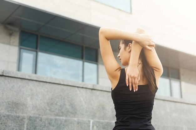 Fitness woman stretching her back before running outdoors, back view. Young slim girl makes aerobics exercise, gray wall background, copy space