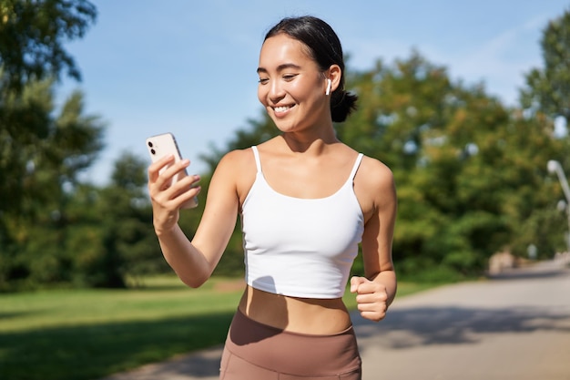 Fitness woman with water bottle and smartphone jogging in park and smiling looking at her mobile pho