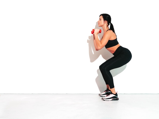 Fitness zelfverzekerde vrouw in zwarte sportkleding Sexy jonge mooie model met perfect lichaam vrouw geïsoleerd op een witte muur in de studio die zich uitstrekt voor de trainingSquats maken met halters