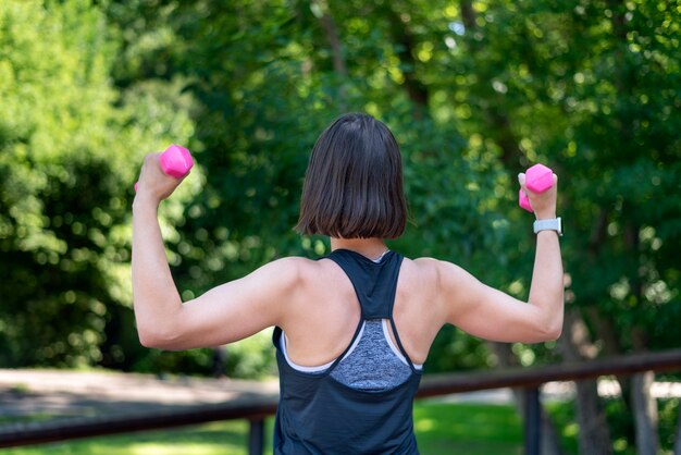 Fitnesstrainer voert training in het park. Meisje houdt kleine halters vast en doet de oefening. Achteraanzicht.