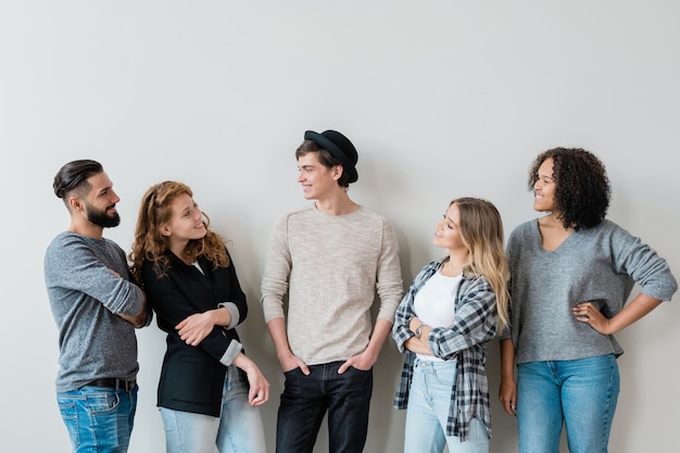 Five casual carefree guys and girls having friendly talk at leisure while standing against white wall in studio