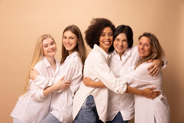 Five female friends of different races and ages with sincere smiles look at camera hugging each other on beige background