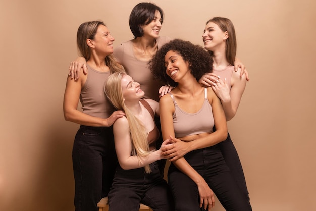 Five happy interracial ladies of different ages in brown tops and black pants smile at each other on beige background