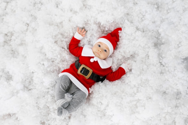 A five month old baby in a Santa suit lies on his back in the snow and smiles