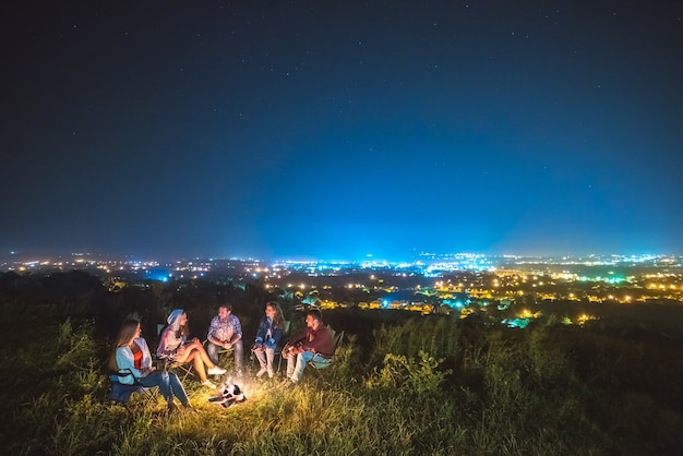 The five people sit near the bonfire on the background of the city. night time