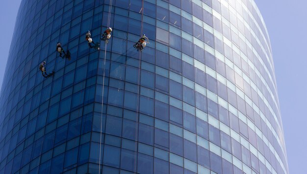 Five window washers hanging with ropes outside blue glass modern building Risky job dangerous work