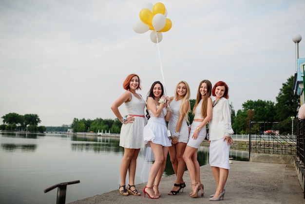 Five women with balloons at hand weared on white dresses on hen party against pier on lake.