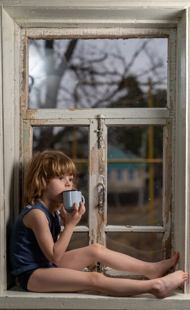 A five-year-old boy with a sad look sits on a shabby wooden windowsill with a metal mug in his hands