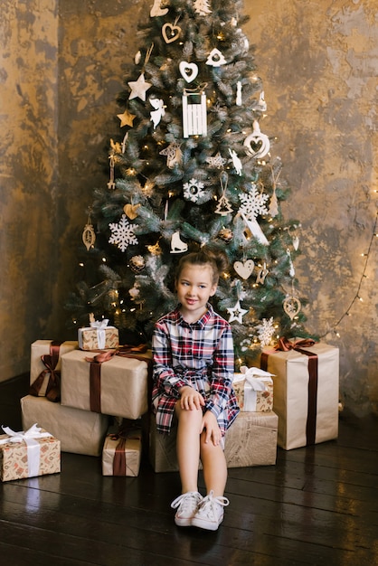 A five-year-old girl in a plaid dress sits near a Christmas tree on new year's gifts and smiles. She is happy and contented