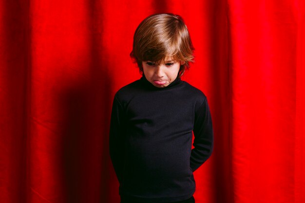 Five years old sad boy, wearing black clothes, against a red curtain