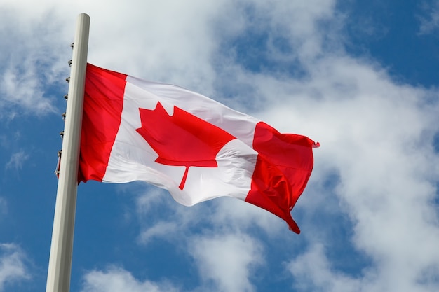 Flag of Canada on a flagpole flutters in the wind against a bright blue sky with clouds