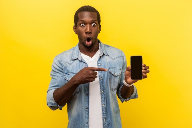 Flagman-gadget. Portret van een verbaasde man in een spijkerblouse die naar de mobiele telefoon wijst en met grote ogen naar de camera kijkt en zegt wow verbaasd over technologie. indoor studio-opname geïsoleerd op gele achtergrond