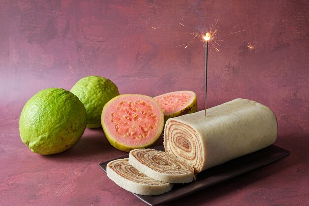 Flame of a candle sparkles on a cake roll next to guavas.