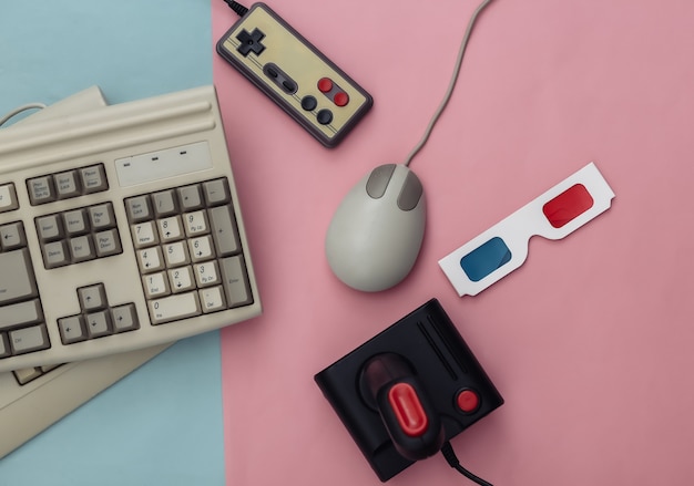 フラットレイ80年代の属性構成。 PCキーボード、マウス、ゲームパッド、3Dメガネ。レトロな電子機器、ガジェット、デバイス。エンターテインメントとゲーム。ピンクブルーの背景。上面図