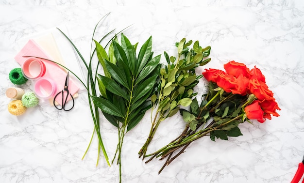 Flat lay. Arranging red roses and green leaves into a bouquet on a marble background.