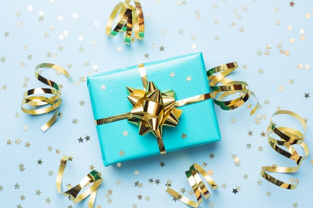 Flat lay background for celebration Christmas and New Year. Gift box turquoise with gold ribbons bows and confetti stars on a blue background. top view copy space.