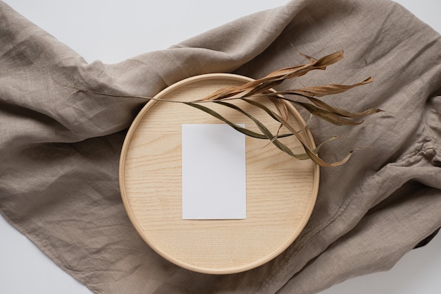 Flat lay of blank paper business card with flowers on wooden tray on neutral beige blanket