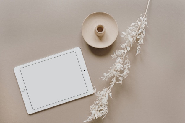 Flat lay of blank screen tablet pad, white floral branch, decorations on neutral pastel beige. Home office desk workspace. Flat lay, top view.