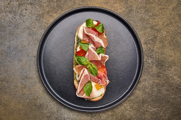 Flat lay bruschetta with mozzarella parma ham and pesto on black plate on textured background