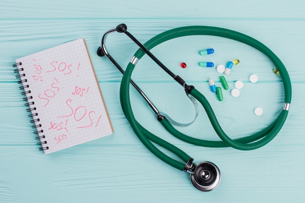 Flat lay capsules and stethoscope on blue background. Sos written on notepad.