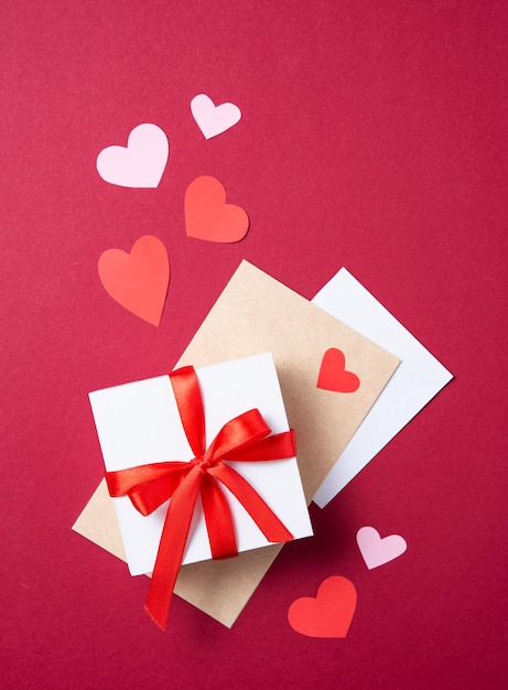 Flat lay of celebration. White gift box, and craft letter on a red background with papper hearts. Concept of Valentine's Day, Mother's Day, wedding day. Top view and copy space