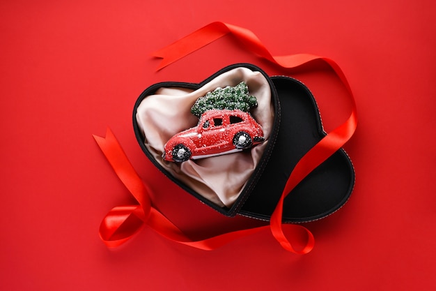 Flat lay Christmas composition. Small red toy car in a black box in the form of a heart with a red ribbon on red.