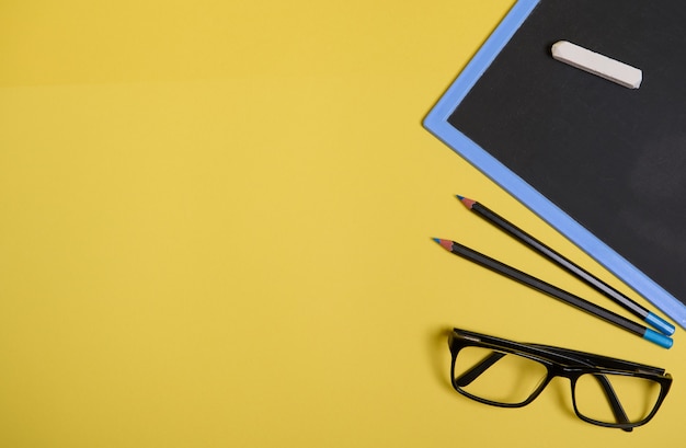 Flat lay composition. Chalk on a clean blackboard with copy space , eyeglasses and two pencils, isolated on yellow background with space for text