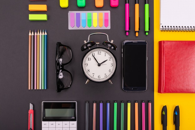 Фото Плоская композиция из делового стола с будильником, смартфоном, ноутбуком, наклейками и цветными ручками на черно-желтом цвете