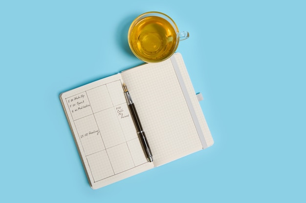 Photo flat lay composition of an open agenda with schedule, ink pen and transparent glass cup of tea lie on blue surface. color background with copy space . time management, deadline, schedule concept