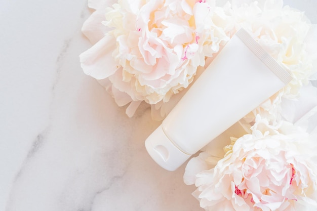 Flat lay composition with cosmetic products and peony flowers on a marble Top view Unbranded plastic white tube with face or hand cream or cosmetic mask