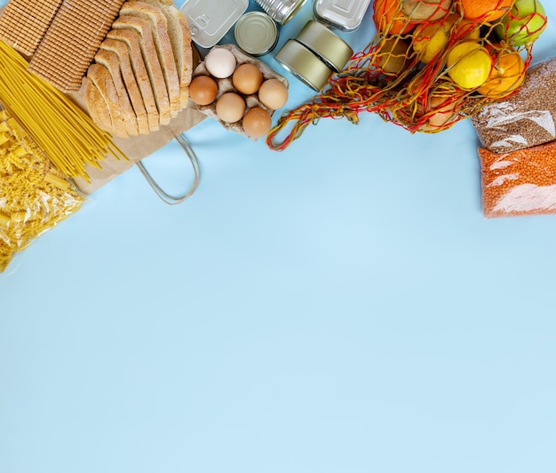 Плоская композиция с фруктами, яйцами, хлебом, печеньем, макаронами, чечевицей, консервами на синем фоне. Место для текста.