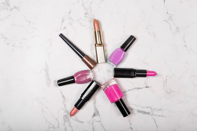 Flat lay of cosmetic makeup products including lipsticks, nail varnish on white marble background.