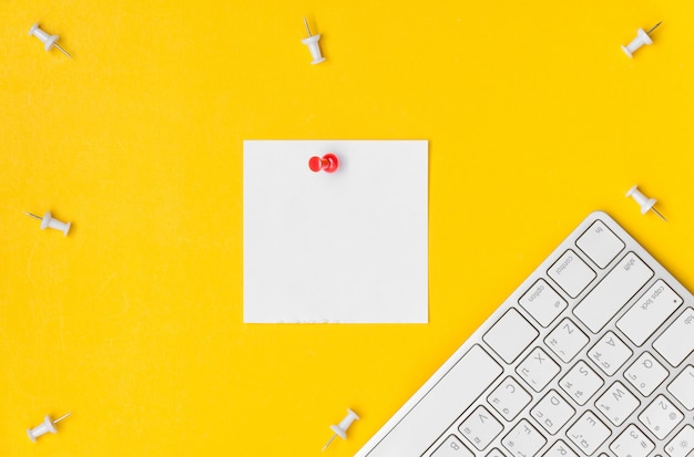 Flat lay of desktop workspace with red pushpin sticky on blank space white paper noted and keyboard on yellow background