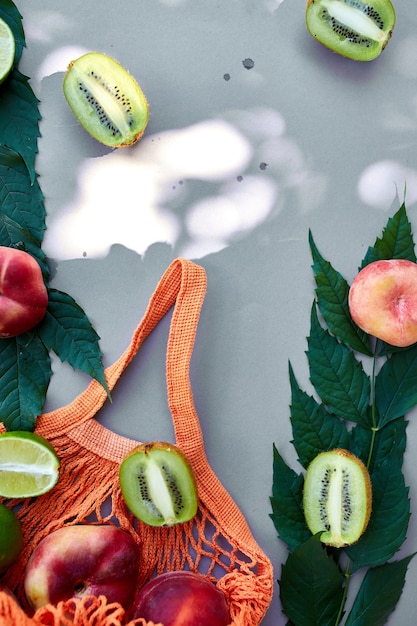 Foto lay piatto di eco-friendly mesh shopping bag con frutta pesca, kiwi, lime su sfondo grigio alla luce del sole, ora legale. concetto di drogheria, copia spazio, vista dall'alto.
