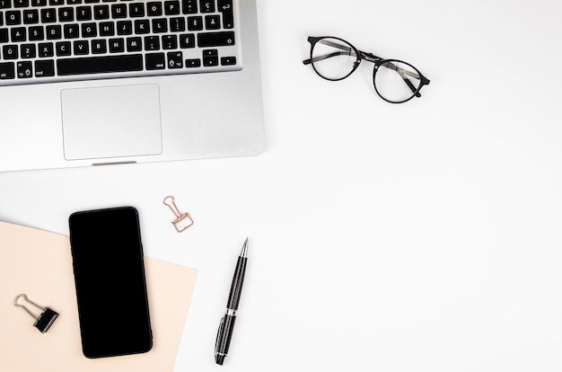 Flat lay frame mockup white Desk with a silver laptop, glasses, pen and smartphone with a black screen