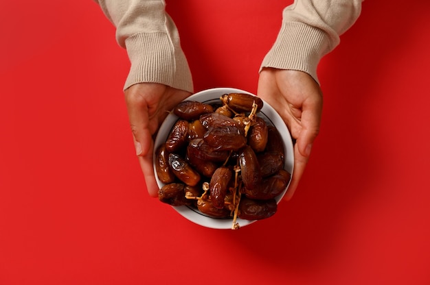 Photo flat lay . hands holding a delicious and sweet plate full of fresh dates, isolated on red background with copy space