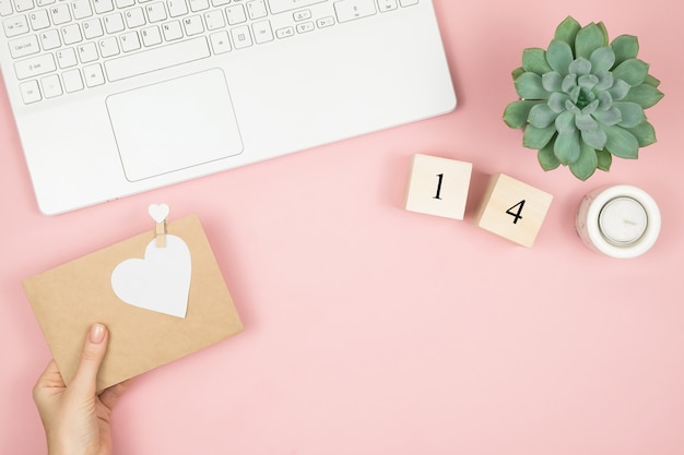 Flat lay home office desk. Women workspace with laptop, cosmetics, perfume, accessories on pink surface. Love letter envelope, valentine red heart