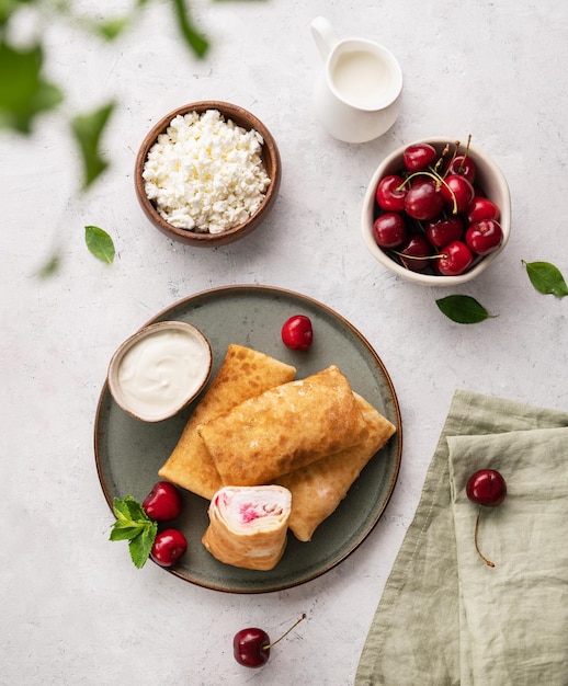 Flat lay homemade pancakes or crepes with cottage cheese and fresh cherries on a green plate with sour cream on a light background Top view