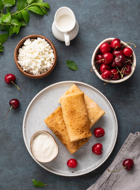 Flat lay homemade pancakes or crepes with cottage cheese and fresh cherries on a plate with sour cream on a dark blue background Top view